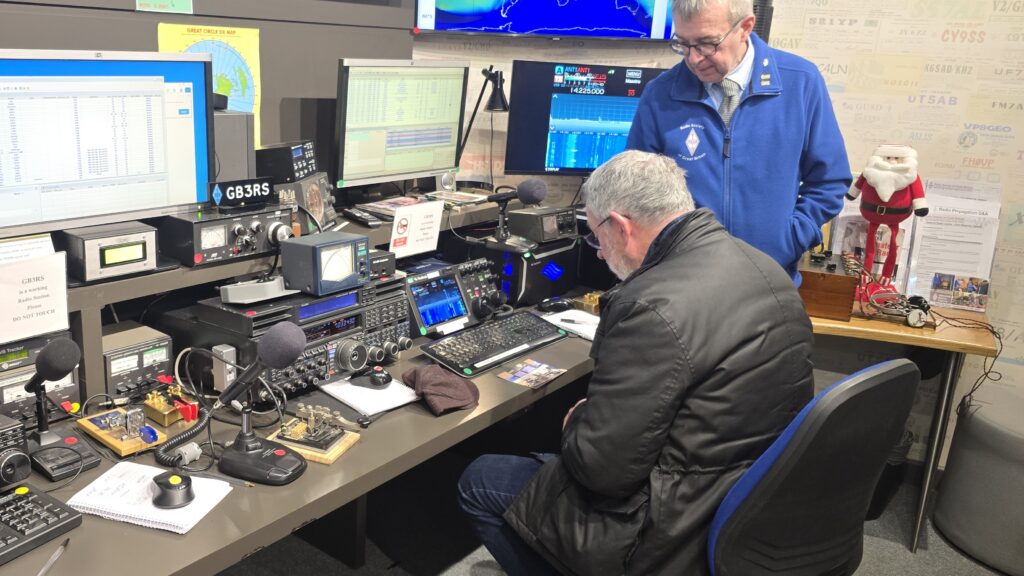 An Amateur Radio operator - Peter G4ENC - sitting at a table in front of the RSGB's radio station GB3RS. Also present is Simon MØKBJ - a National Radio Centre volunteer.